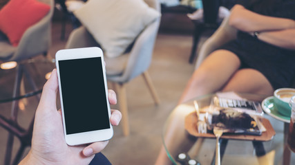 Mockup image of a hand holding white mobile phone with blank black screen in modern cafe and blur Asian woman in background