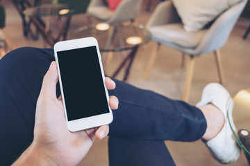 Mockup image of a man's hand holding white mobile phone with blank black screen on thigh with white canvas shoes in cafe with feeling relax