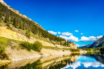 Fototapeta na wymiar Reflections of blue sky, trees and mountains in the smooth surface on the crystal clear water of Crown Lake, along Highway 99 in Marble Canyon Provincial Park in British Columbia, Canada