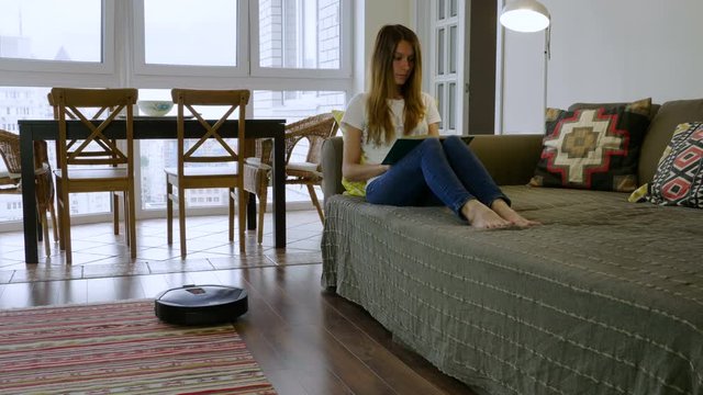 The young beautiful woman reading a book on the sofa while the robot cleaner cleaning the floor in the room. 4K