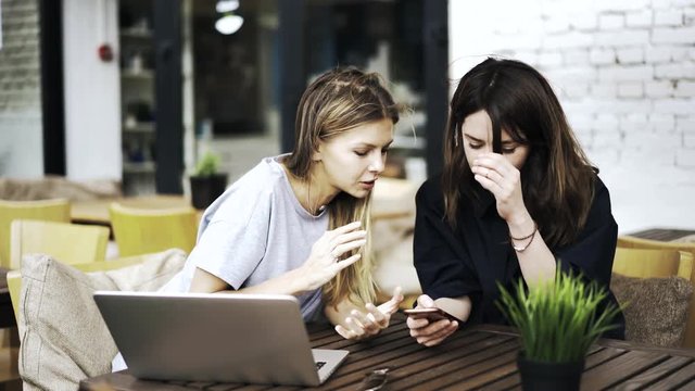 Two young women are sitting together in a cafe and discussing their lives while looking at a smartphone screen and smiling. Concept of good times. Locked down real time medium shot