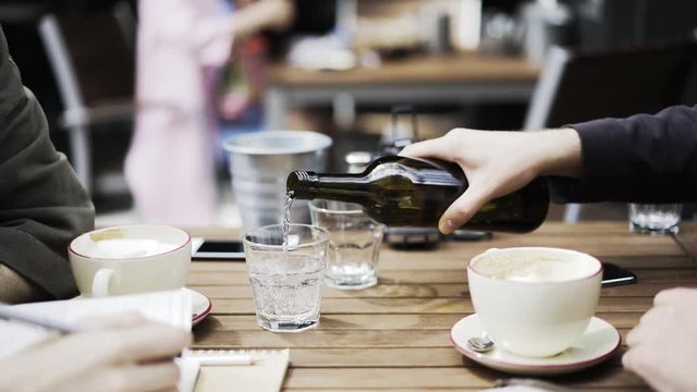 Hand of an unrecognizable young man pouring some mineral water into his friends glass while they are talking in an outdoors cafe. Locked down real time medium shot