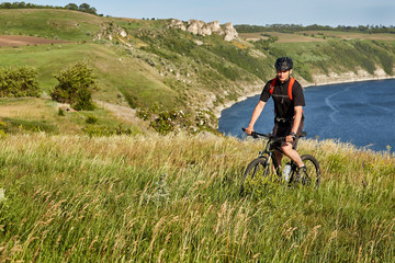 Young man riding mountain bike on the green meadow above the blue river in the countryside.