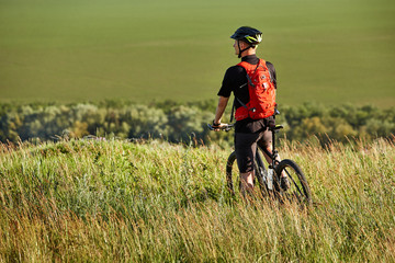 Obraz premium Rear view of the sportsman with his mountain bike stands on the meadow and looking away in the countryside.