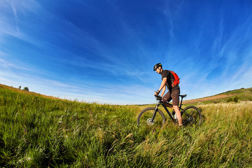 Young cyclist cycling on the green meadow against beautiful blue sky with clouds in the countryside.