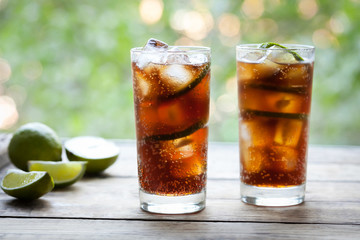 Cuba Libre refreshing summer cocktail with a cola, ice, lime and rum on wooden table with a view to the terrace and trees. Close up summer beverage. Cold longdrink or lemonade.A frozen glass