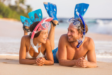 Summer travel beach vacation snorkel couple talking together laughing having fun relaxing lying down on sand at sunset. Girl, man wearing diving mask and fins happy. - Powered by Adobe