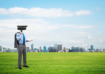 Camera headed man standing on green grass against modern cityscape