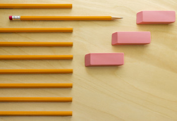 Pattern of pencils and erasers on light wood desk, one pencil sharpened and standing out