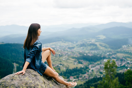 Lonely young traveler girl siiting on stone at top of mountain. Discovering new country. Beautiful nature landscape from high altitude. Summer trip on weekend vacation.  Edge of world. Jeans clothes.