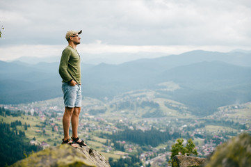 Lonely traveler man in summer clothes standing on stone at top of carpathian mountain and looking far from high altitude. Nature landscape with far horizon. Tourist resting in wild terrain on vacation