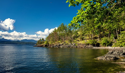 Landscape of the northern sea against the backdrop of a wooded slope, range of snow-capped mountains and a cloudy sky.