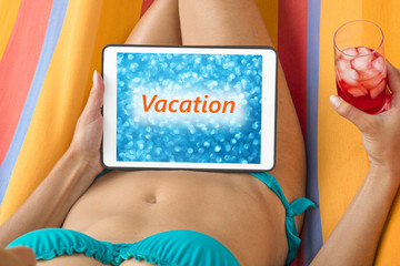 Young woman in bikini on a hammock with tablet device looking at blurred blue background with word "Vacation" written on it