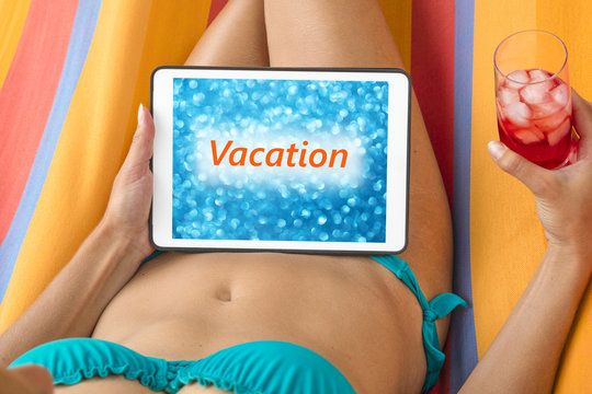 Young woman in bikini on a hammock with tablet device looking at blurred blue background with word "Vacation" written on it