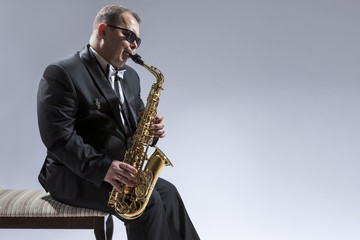 Fototapeta na wymiar Music Concepts. Portrait of Mature Relaxed and Thoughful Caucasian Saxophone Player in Sunglasses Playing the Saxophone While Sitting on Chair in Studio Environment.