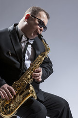 Fototapeta na wymiar Male Mature Saxophone Player in Sunglasses Playing the Saxophone While Sitting on Chair in Studio Environment.