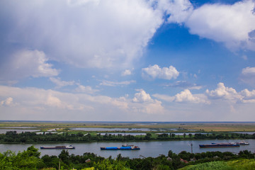 Fototapeta premium Summer landscape overlooking the river. Boats sail along the picturesque edge. The barge floats on the river. Quaint clouds in the sky.