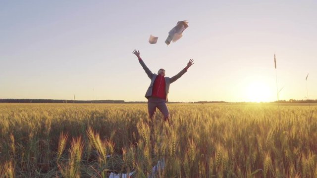 Businessman in a suit on a wheat field throws a stack of documents up and triumphantly throws up his hands, slow motion