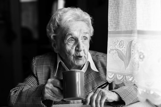 Black-and-white portrait of an elderly woman drink tea near the window in house