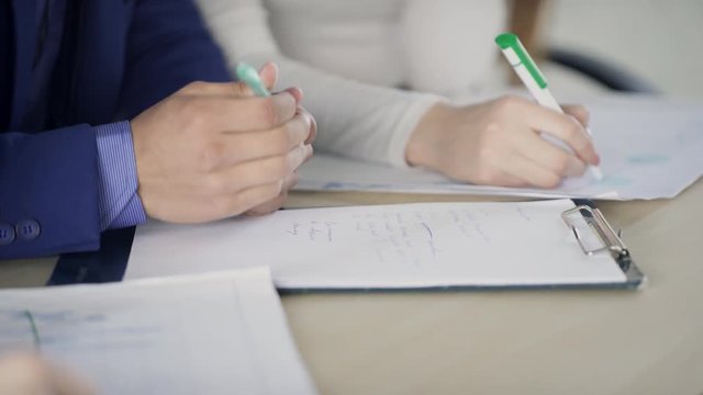 Male and female hands are writing on paper at the desk. The chief in a blue business suit shows the secretary, who keeps the record, about important issues that need to be reflected in the documents