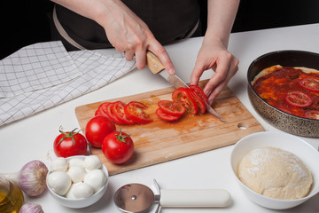Female chef cuts the tomatoes for making homemade pizza. On the white table are mozzarella, pizza dough, and garlic