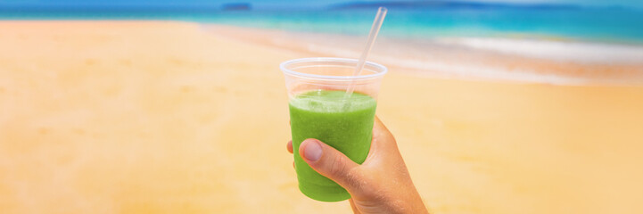 Green smoothie healthy juice detox drink banner. Food selfie man holding plastic cup on beach summer background. Panorama crop for copy space.