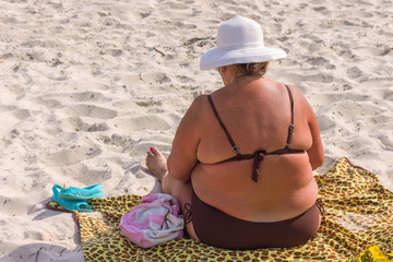 an overweight woman in white hat sitting on a beach back view