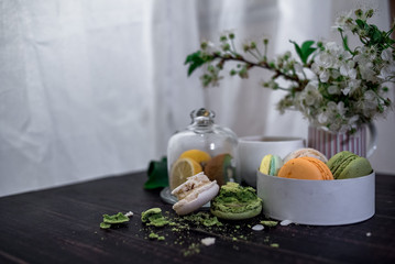 Crushed pastel, orange and green macaroons on a wood background with whole one in white plate with jug with flowers on the back