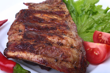 Pork ribs on grill and chili pepper with tomato, lettuce leaves on white plate