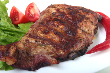 Pork ribs on grill and chili pepper with tomato, lettuce leaves on white plate