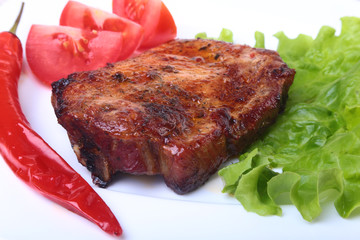 Steak of grilled meat and chili pepper with tomato, lettuce leaves on white plate.