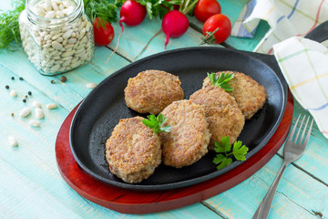 Homemade meatballs with white beans and fresh herbs. Cast-iron frying pan full of delicious fried meat cutlets, fresh greens and vegetables on the kitchen table.