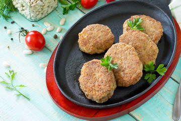 Domestic meat cutlet with white beans and fresh herbs. Cast-iron frying pan full of delicious fried meat cutlets, fresh greens and vegetables on the kitchen table.