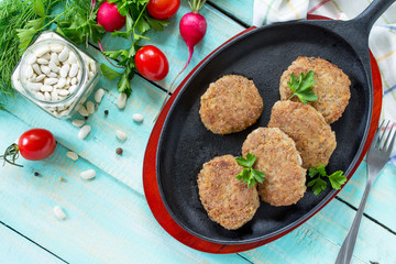 Homemade meatballs with white beans and fresh herbs. Cast-iron frying pan full of delicious fried meat cutlets, fresh greens and vegetables on the kitchen table. Top view.