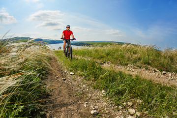 Young cyclist riding the mountain bike on the beautiful summer trail in the countryside against blue sky with clouds.