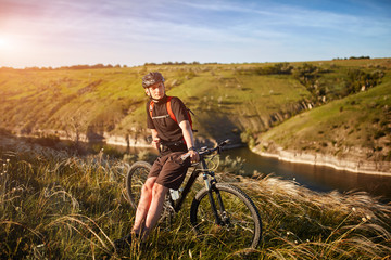 Obraz premium Portrait of the young cyclist standing on the hill above the river against blue sky with clouds.