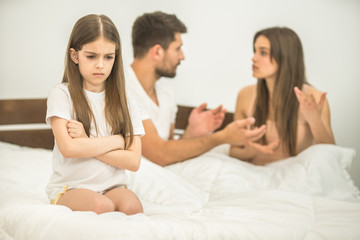 The sad girl sitting near the parents on the bed