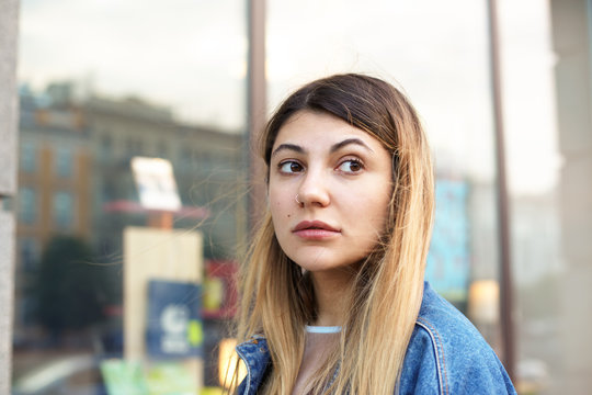 Attractive mixed race young woman with long bleached hair, brown eyes and facial piercing, standing on city streets, waiting for friends for a walk, looking aside with serious expression on her face