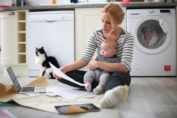 Woman holding baby son in arms, looking through work on kitchen floor, laptop and digital tablet in front of her