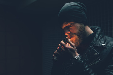 Young male hipster in knit hat igniting cigarette at night