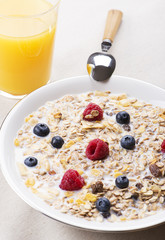 Cereal bowl breakfast with milk, raspberries and blueberries next to orange juice. Vertical studio shot.