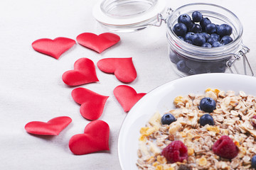 Breakfast cereals with raspberries and blueberries next to hearts on cloth tablecloth.