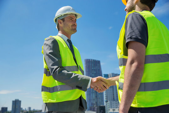 Two men wearing hard hats and hi vis vests, shaking hands, outdoors