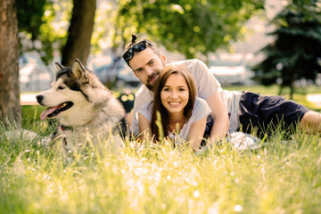 Handsome beard man with beautiful young woman and their dog alaskan malamute lying on green grass in summer time outdoor. Couple lying on the grass with a dog in the park.