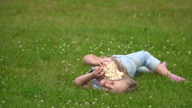 A bit the fair-haired girl listens to a surf of waves in a big cockleshell. The beautiful little girl lies on a green glade with a cockleshell in a hand