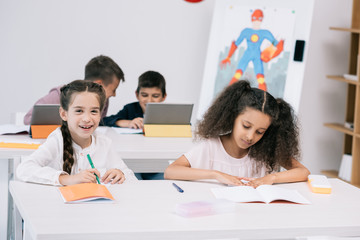 Multiethnic group of pupils writing in exercise books and using digital tablets during lesson