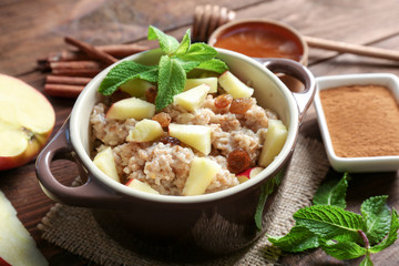 Tureen with tasty oatmeal porridge, apples, raisins and cinnamon on wooden table, closeup