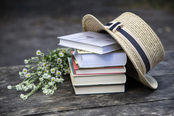 A stack of books on an old wooden table next to a straw hat of a canoe and a bouquet of wild flowers