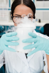 Young female scientist in eyeglasses holding flask while making experiment in chemical lab