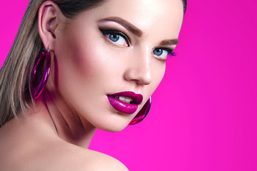 A beautiful, sweet, well-groomed girl on a bright pink background with combed smooth hair and large pink shiny earrings. Makeup - pink lipstick, black arrows. Blue eyes. Fashion, luxury, cosmetics.